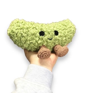 Könnte beinhalten: Eine grüne Plüschbohne mit einem lächelnden Gesicht und zwei braunen Beinen. Die Bohne ist aus flauschigem Stoff und hat schwarze Augen.