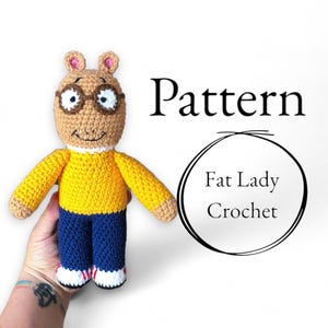 Puede incluir: Muñeco de Arthur tejido a ganchillo con un suéter amarillo, pantalones azules y gafas. El muñeco tiene una cabeza color canela con orejas marrones y lo sostiene una persona. La imagen incluye el texto "Pattern" y "Fat Lady Crochet."