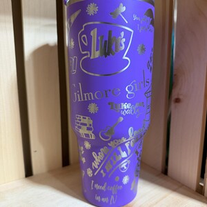 Pu&ograve; includere: Un bicchiere termico viola con coperchio argentato e disegni incisi argentati. Il bicchiere presenta il testo "Luke's" all'interno di una tazza di caff&egrave;, insieme ad altri testi e grafiche. Sono visibili anche le parole "Gilmore girls" e "I need coffee in an N".