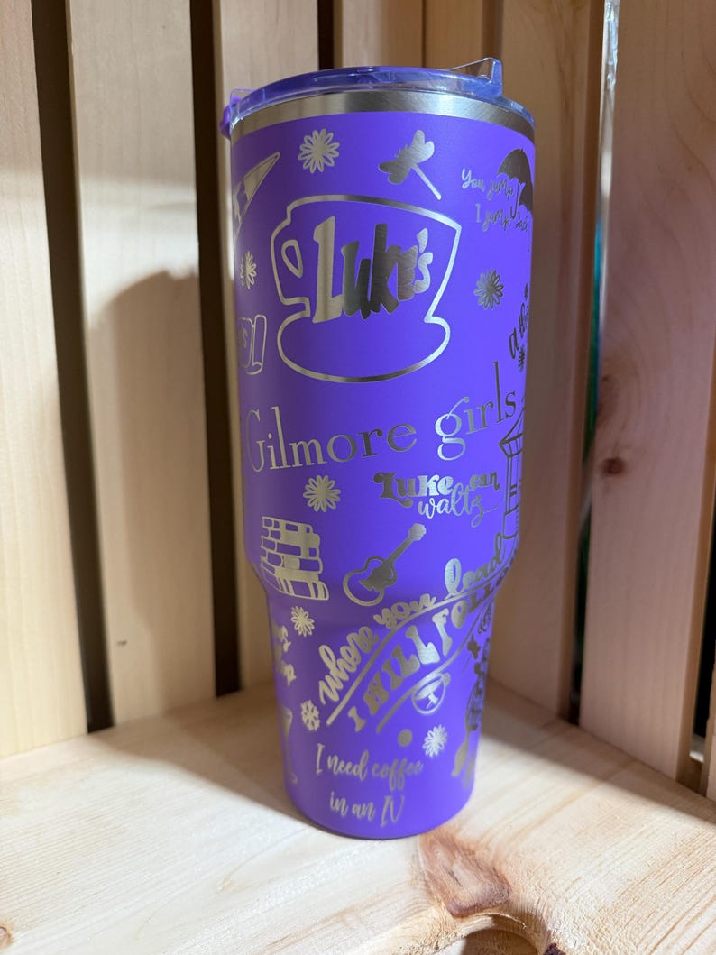 Pu&ograve; includere: Un bicchiere termico viola con coperchio argentato e disegni incisi argentati. Il bicchiere presenta il testo "Luke's" all'interno di una tazza di caff&egrave;, insieme ad altri testi e grafiche. Sono visibili anche le parole "Gilmore girls" e "I need coffee in an N".