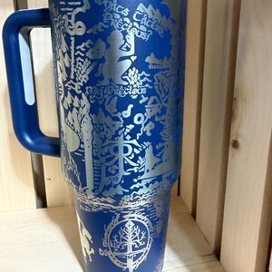 May include: A blue stainless steel tumbler with a handle and a clear lid. The tumbler features an engraved design with text and illustrations. The design includes the text "What's precious?" and a tree with a sword.