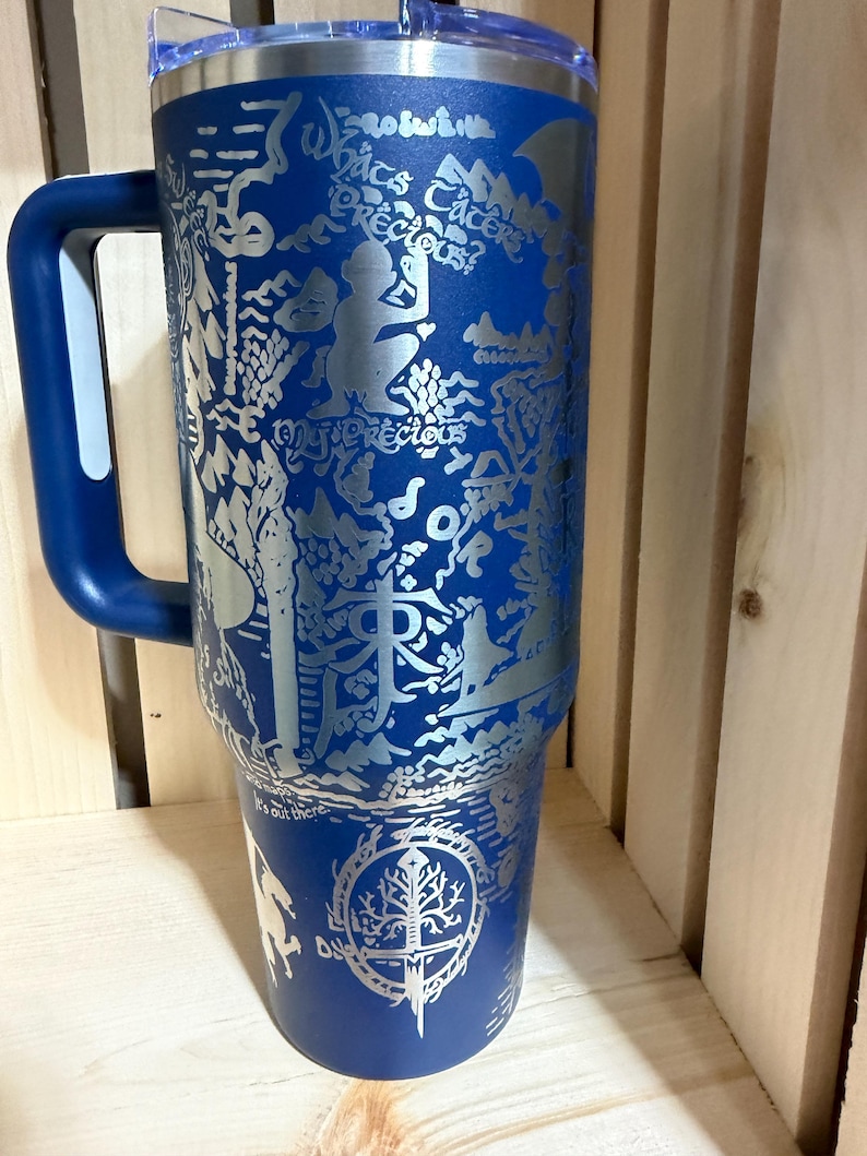May include: A blue stainless steel tumbler with a handle and a clear lid. The tumbler features an engraved design with text and illustrations. The design includes the text "What's precious?" and a tree with a sword.