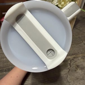 May include: A white and gray plastic lid for a container. The lid has a gray handle and a gray bar across the center. The lid is round and has a light blue interior.
