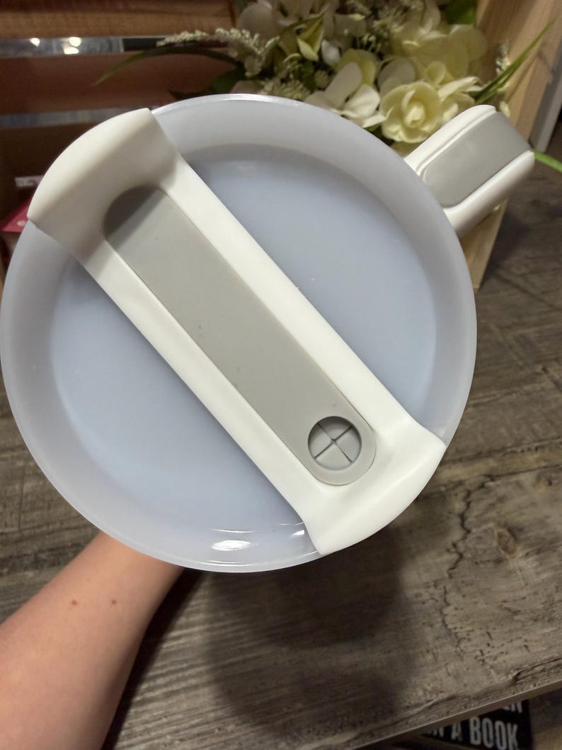 May include: A white and gray plastic lid for a container. The lid has a gray handle and a gray bar across the center. The lid is round and has a light blue interior.