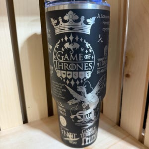 Pu&ograve; includere: Bicchiere in acciaio inossidabile nero con un design argentato. Presenta le parole "Game of Thrones", citazioni e simboli. Il design include una corona, un drago e altre immagini della serie. Il bicchiere ha un coperchio trasparente.