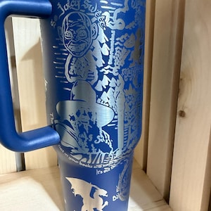 May include: A blue stainless steel tumbler with a handle, featuring etched illustrations and text. The design includes a cartoon face, a figure in a hat, a dragon, and the words "So Juicy Sweet" and "Not in your books and maps, it's out there!"
