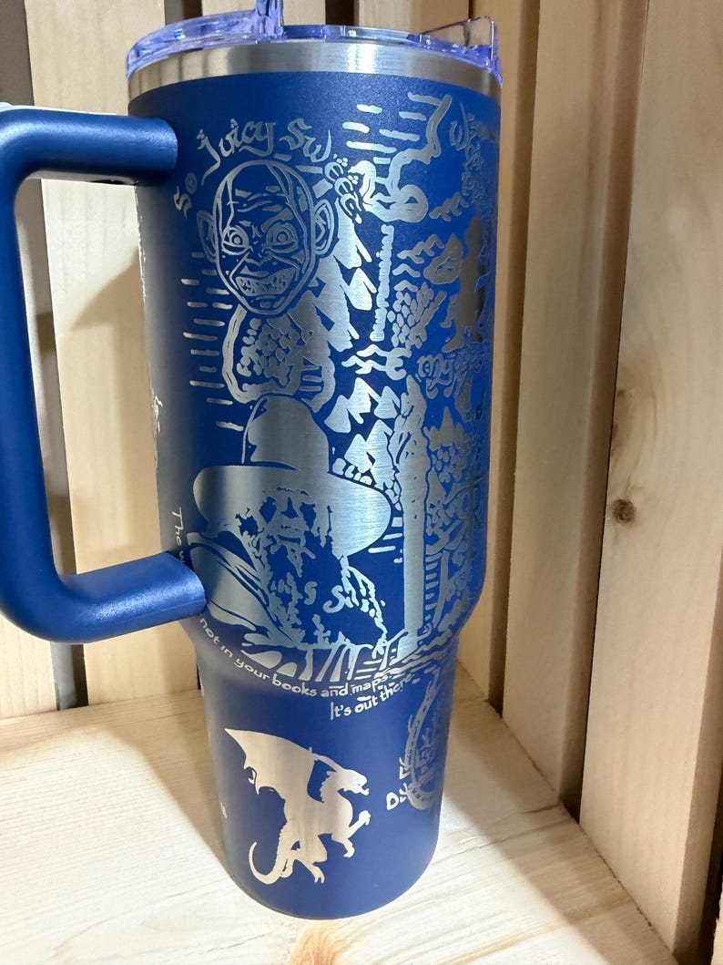 May include: A blue stainless steel tumbler with a handle, featuring etched illustrations and text. The design includes a cartoon face, a figure in a hat, a dragon, and the words "So Juicy Sweet" and "Not in your books and maps, it's out there!"