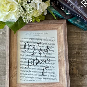 May include: Framed artwork featuring a page from "A Court of Wings and Ruin" with the quote "Only you can decide what breaks you" in a handwritten style. The frame is a light brown wood, and the background includes books and flowers.