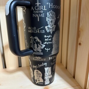 Pu&ograve; includere: Bicchiere in acciaio inossidabile nero con manico, con citazioni e immagini di una serie fantasy. Il bicchiere include frasi come "A Girl Has No Name" e "You Know Nothing Jon Snow."