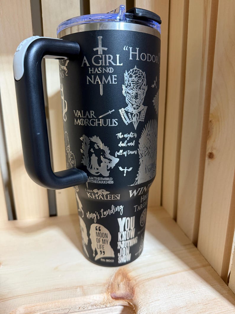 Pu&ograve; includere: Bicchiere in acciaio inossidabile nero con manico, con citazioni e immagini di una serie fantasy. Il bicchiere include frasi come "A Girl Has No Name" e "You Know Nothing Jon Snow."