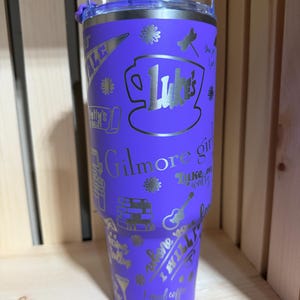 Pu&ograve; includere: Un bicchiere viola in acciaio inossidabile con coperchio argentato. Il bicchiere &egrave; ricoperto di testo e grafica argentati, tra cui le parole "Luke's", "Gilmore Girls" e altre immagini correlate. Il bicchiere &egrave; alto circa 23 cm.