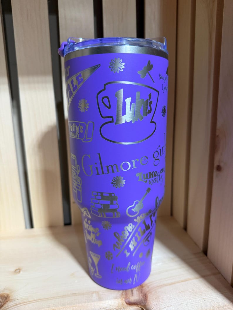 Pu&ograve; includere: Un bicchiere viola in acciaio inossidabile con coperchio argentato. Il bicchiere &egrave; ricoperto di testo e grafica argentati, tra cui le parole "Luke's", "Gilmore Girls" e altre immagini correlate. Il bicchiere &egrave; alto circa 23 cm.