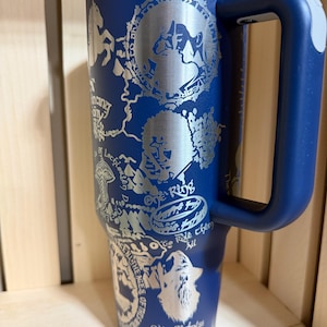 May include: A navy blue insulated tumbler with a handle and a silver lid. The tumbler features engraved illustrations and text, including "One Ring" and "Eden Darkness Music Pass."