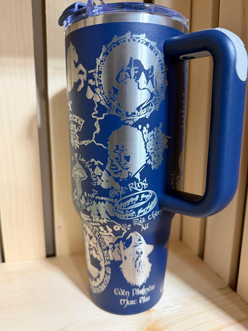 May include: A navy blue insulated tumbler with a handle and a silver lid. The tumbler features engraved illustrations and text, including "One Ring" and "Eden Darkness Music Pass."