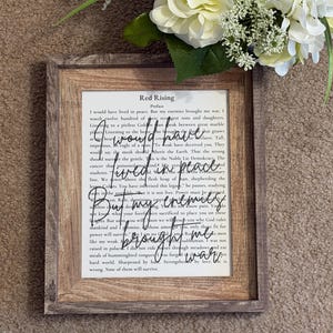 May include: Framed print of a book page with the title "Red Rising" and handwritten text overlay. The frame is a light brown color. White flowers are in the background.