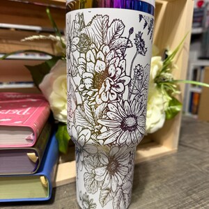 May include: White insulated tumbler with a floral design. The tumbler features a white lid and a band of iridescent color near the top. The floral pattern includes daisies and other flowers in a black outline.