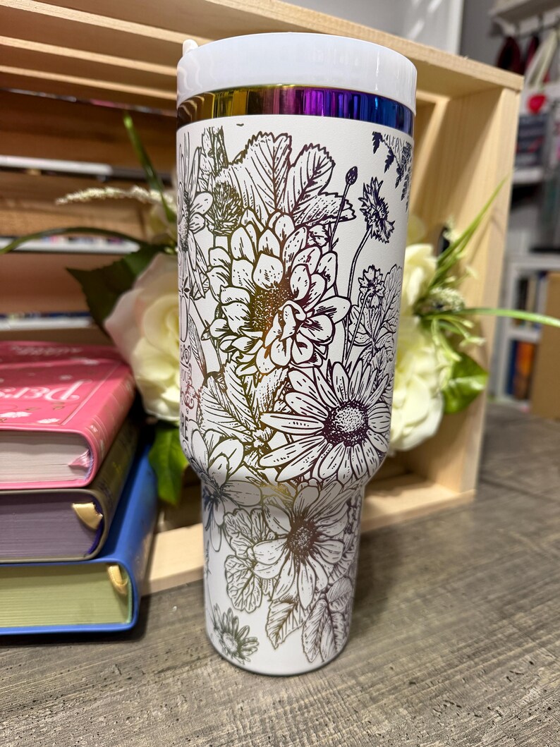 May include: White insulated tumbler with a floral design. The tumbler features a white lid and a band of iridescent color near the top. The floral pattern includes daisies and other flowers in a black outline.