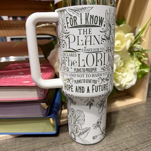 May include: A white travel mug with a white handle and lid. The mug features a floral design and the text "FOR I KNOW THE PLANS I HAVE FOR YOU...HOPE AND A FUTURE" from Jeremiah 29:11. The mug is next to books and flowers.