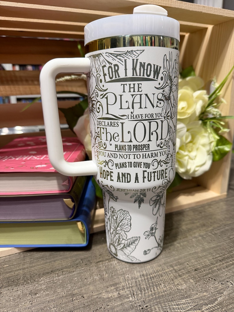 May include: A white travel mug with a white handle and lid. The mug features a floral design and the text "FOR I KNOW THE PLANS I HAVE FOR YOU...HOPE AND A FUTURE" from Jeremiah 29:11. The mug is next to books and flowers.