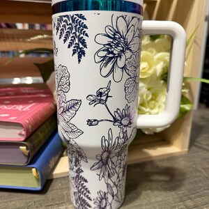 May include: White insulated tumbler with a large white handle and lid. The tumbler is decorated with a floral design in navy blue. The top of the tumbler has a teal-colored band.