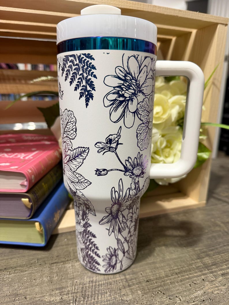 May include: White insulated tumbler with a large white handle and lid. The tumbler is decorated with a floral design in navy blue. The top of the tumbler has a teal-colored band.