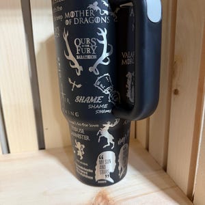 Pu&ograve; includere: Bicchiere termico nero con manico e coperchio argentato. Il bicchiere presenta disegni e frasi incise, tra cui "Mother of Dragons", "Ours is the Fury" e "My Sun and Stars". Il manico &egrave; nero con un'impugnatura grigia.