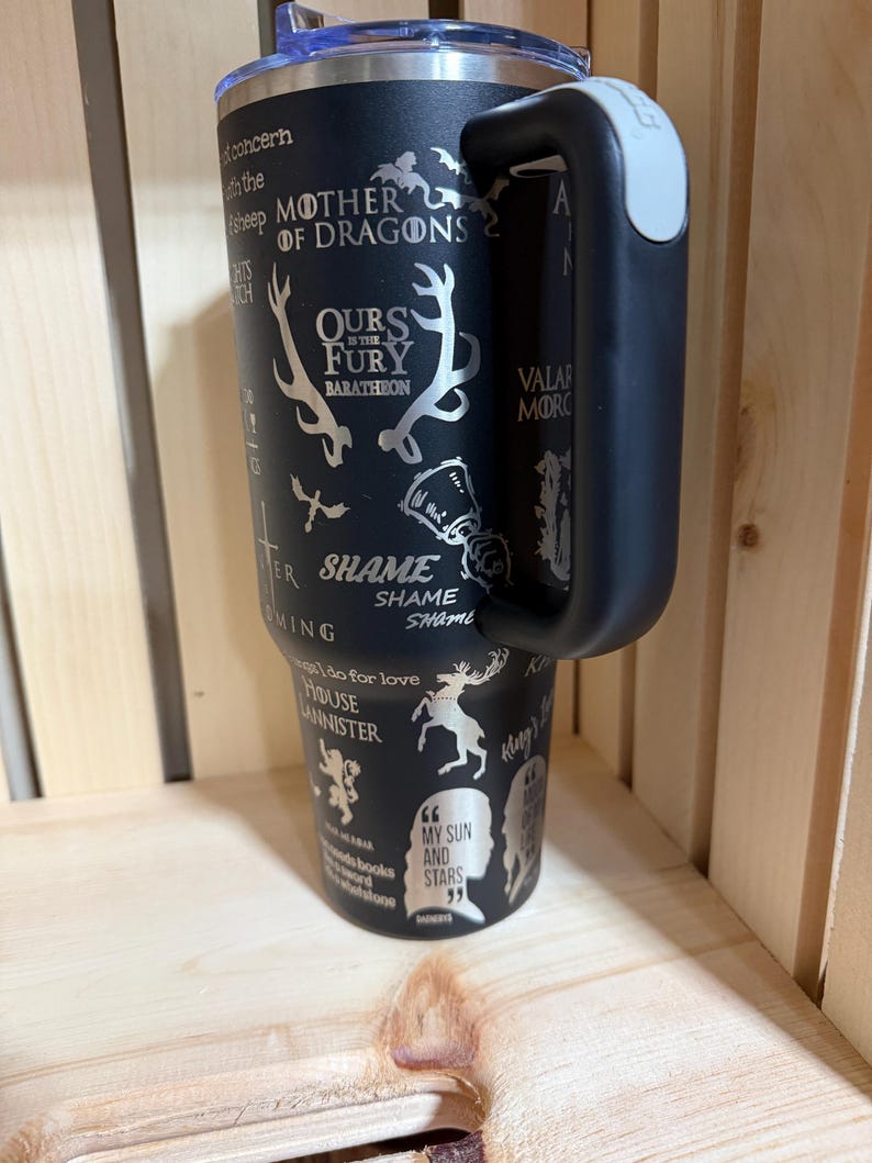 Pu&ograve; includere: Bicchiere termico nero con manico e coperchio argentato. Il bicchiere presenta disegni e frasi incise, tra cui "Mother of Dragons", "Ours is the Fury" e "My Sun and Stars". Il manico &egrave; nero con un'impugnatura grigia.