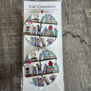 May include: Two car coasters with a bookshelf design. Each coaster features a white background with colorful books, a teacup, a rose in a glass dome, and an apple. The packaging reads "Car Coasters" and "Handmade with love."