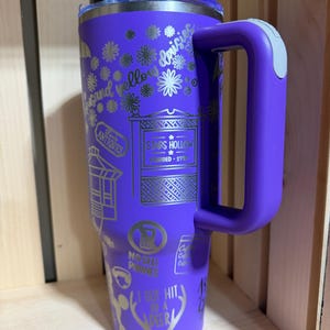 Pu&ograve; includere: Un bicchiere da viaggio viola con manico e coperchio trasparente. La tazza presenta disegni argentati incisi, tra cui fiori, testo e le parole "Stars Hollow". Il manico ha un accento grigio.