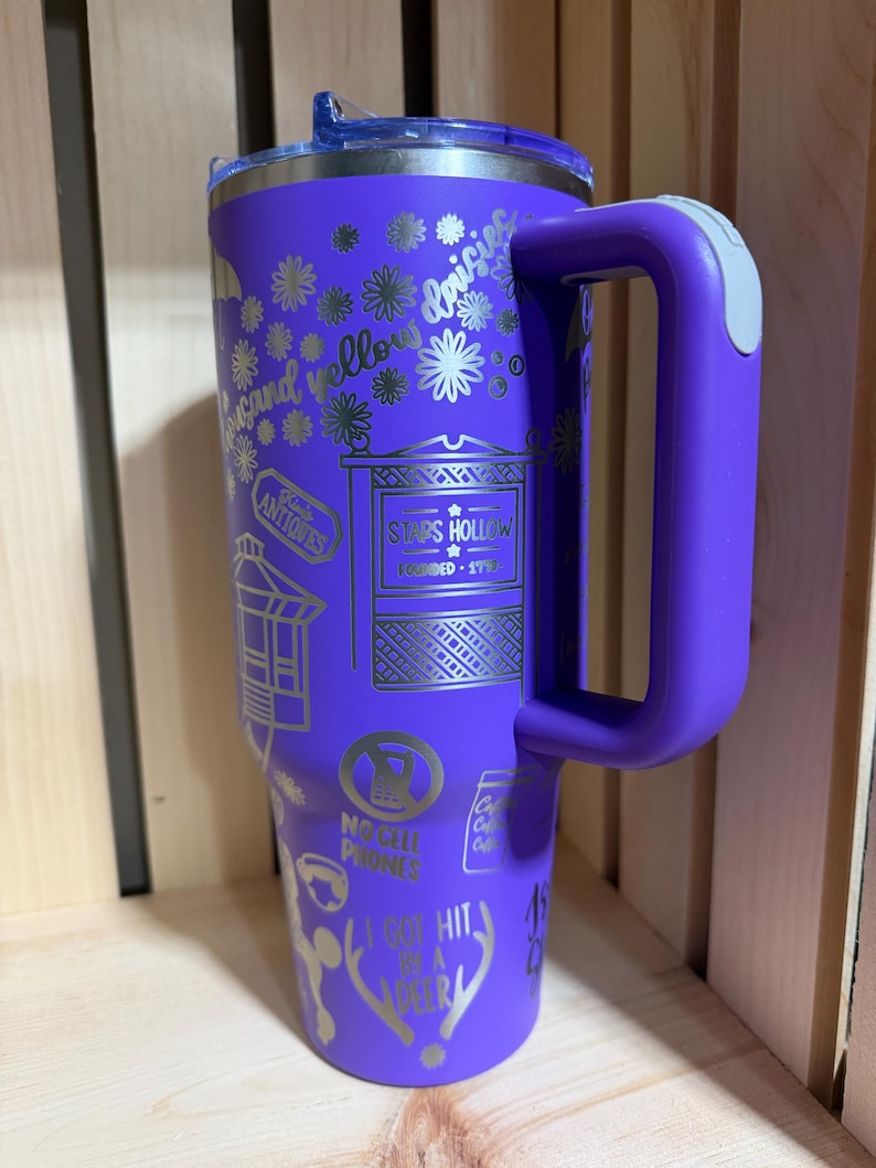 Pu&ograve; includere: Un bicchiere da viaggio viola con manico e coperchio trasparente. La tazza presenta disegni argentati incisi, tra cui fiori, testo e le parole "Stars Hollow". Il manico ha un accento grigio.