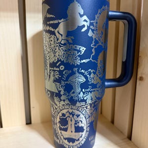 May include: A blue insulated travel mug with a silver design featuring a horse, map, and text. The mug has a handle and a lid. The text includes "Dancing Pony" and other phrases. The mug is designed for hot or cold beverages.
