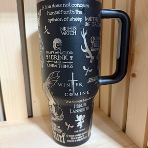 Pu&ograve; includere: Tazza da viaggio isolata nera con manico nero e coperchio blu. La tazza &egrave; ricoperta di citazioni e immagini di una popolare serie fantasy, tra cui le parole "Winter is Coming" e "House Lannister."