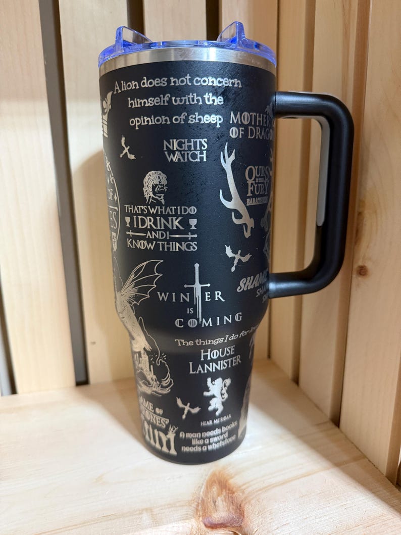 Pu&ograve; includere: Tazza da viaggio isolata nera con manico nero e coperchio blu. La tazza &egrave; ricoperta di citazioni e immagini di una popolare serie fantasy, tra cui le parole "Winter is Coming" e "House Lannister."