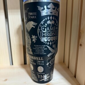 Pu&ograve; includere: Un bicchiere termico nero con coperchio trasparente. Il bicchiere presenta disegni incisi e testo della serie "Game of Thrones", tra cui il sigillo di Casa Stark, le parole "Game of Thrones" e citazioni dello show.
