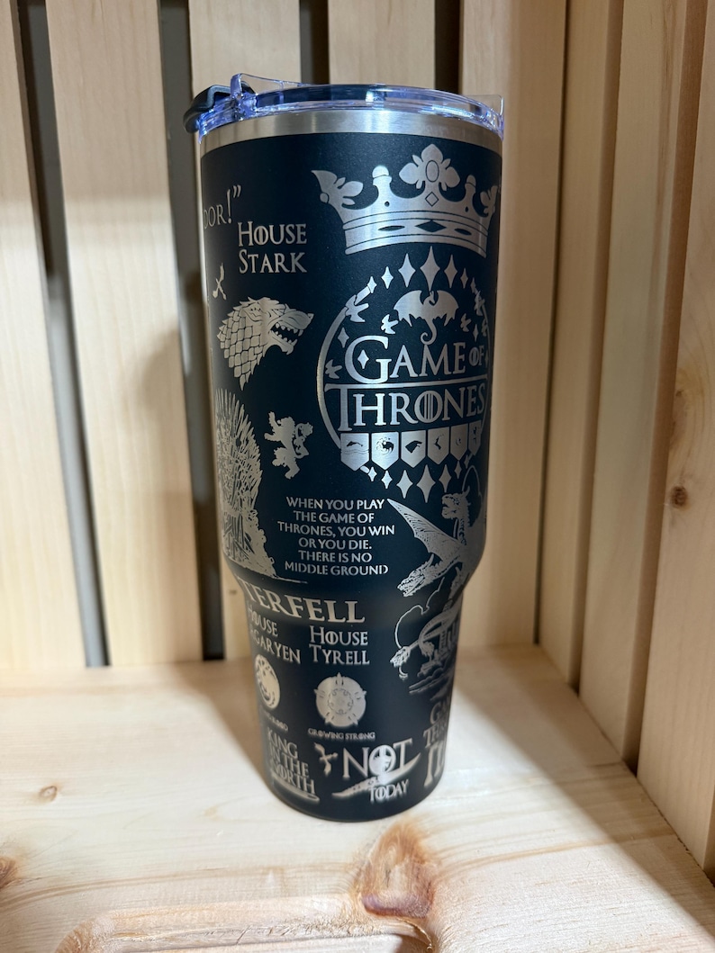Pu&ograve; includere: Un bicchiere termico nero con coperchio trasparente. Il bicchiere presenta disegni incisi e testo della serie "Game of Thrones", tra cui il sigillo di Casa Stark, le parole "Game of Thrones" e citazioni dello show.