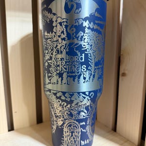 May include: A blue stainless steel tumbler featuring a detailed silver engraving of a map and the words "Lord of the Rings." The design includes a dragon and other imagery from the fantasy series. The tumbler has a clear lid.