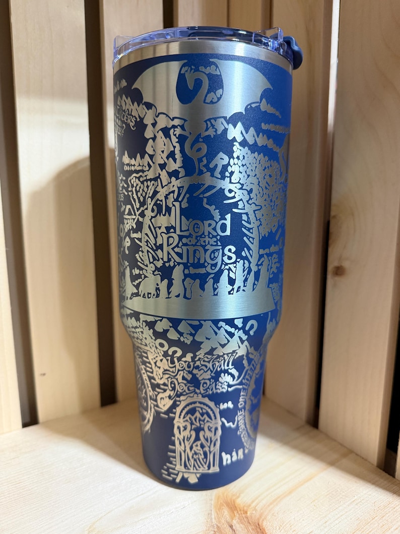 May include: A blue stainless steel tumbler featuring a detailed silver engraving of a map and the words "Lord of the Rings." The design includes a dragon and other imagery from the fantasy series. The tumbler has a clear lid.