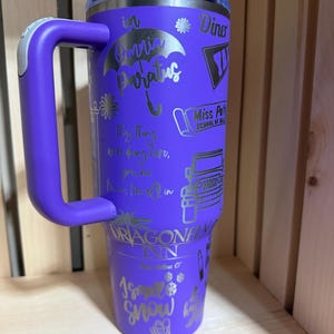 Pu&ograve; includere: Un bicchiere termico viola con manico, con testo e grafica argentati. Il design include frasi come "in Omnia Paratus" e "Dragonfly Inn". Il bicchiere ha un coperchio ed &egrave; posto su uno sfondo di legno.