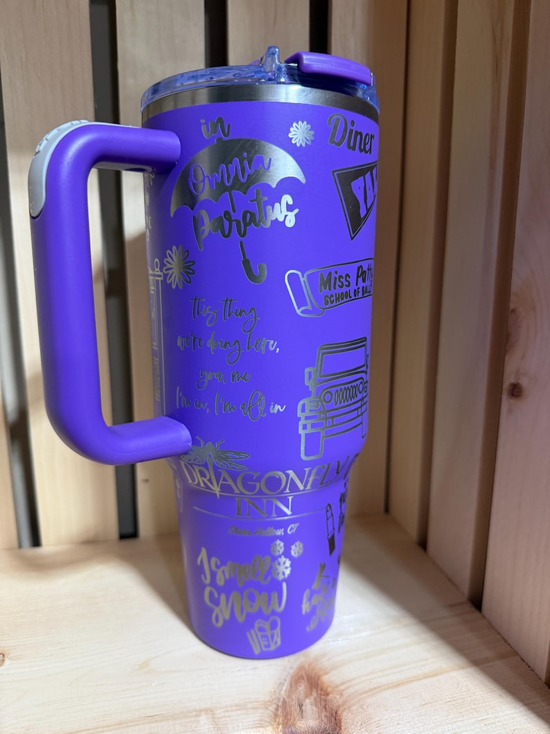 Pu&ograve; includere: Un bicchiere termico viola con manico, con testo e grafica argentati. Il design include frasi come "in Omnia Paratus" e "Dragonfly Inn". Il bicchiere ha un coperchio ed &egrave; posto su uno sfondo di legno.