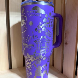 Pu&ograve; includere: Un bicchiere termico viola con manico e coperchio trasparente. Il bicchiere &egrave; decorato con testo e illustrazioni argentate, tra cui le parole "Cafe Girls" e "a thousand yellow". Il design include un barboncino, fiori e un gazebo.