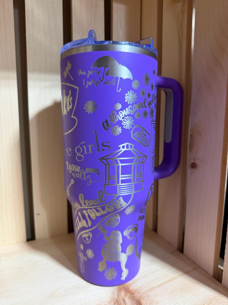 Pu&ograve; includere: Un bicchiere termico viola con manico e coperchio trasparente. Il bicchiere &egrave; decorato con testo e illustrazioni argentate, tra cui le parole "Cafe Girls" e "a thousand yellow". Il design include un barboncino, fiori e un gazebo.