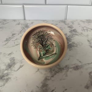 May include: A small, round ceramic bowl with a beige exterior and a swirling interior design of pink, green, and brown. The bowl features a black tree illustration in the center.