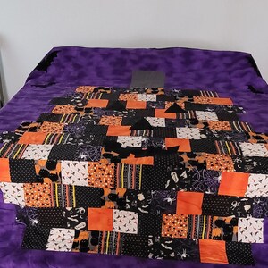 May include: A purple quilt with a patchwork design featuring black, orange, and white fabric. The patchwork is in the shape of a jack-o'-lantern face.