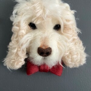 Puede incluir: Una cabeza de perro de fieltro de aguja blanca con una pajarita roja. El perro tiene un pelaje blanco y esponjoso y ojos negros.