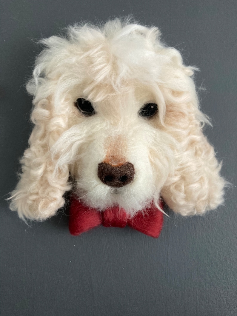 Puede incluir: Una cabeza de perro de fieltro de aguja blanca con una pajarita roja. El perro tiene un pelaje blanco y esponjoso y ojos negros.