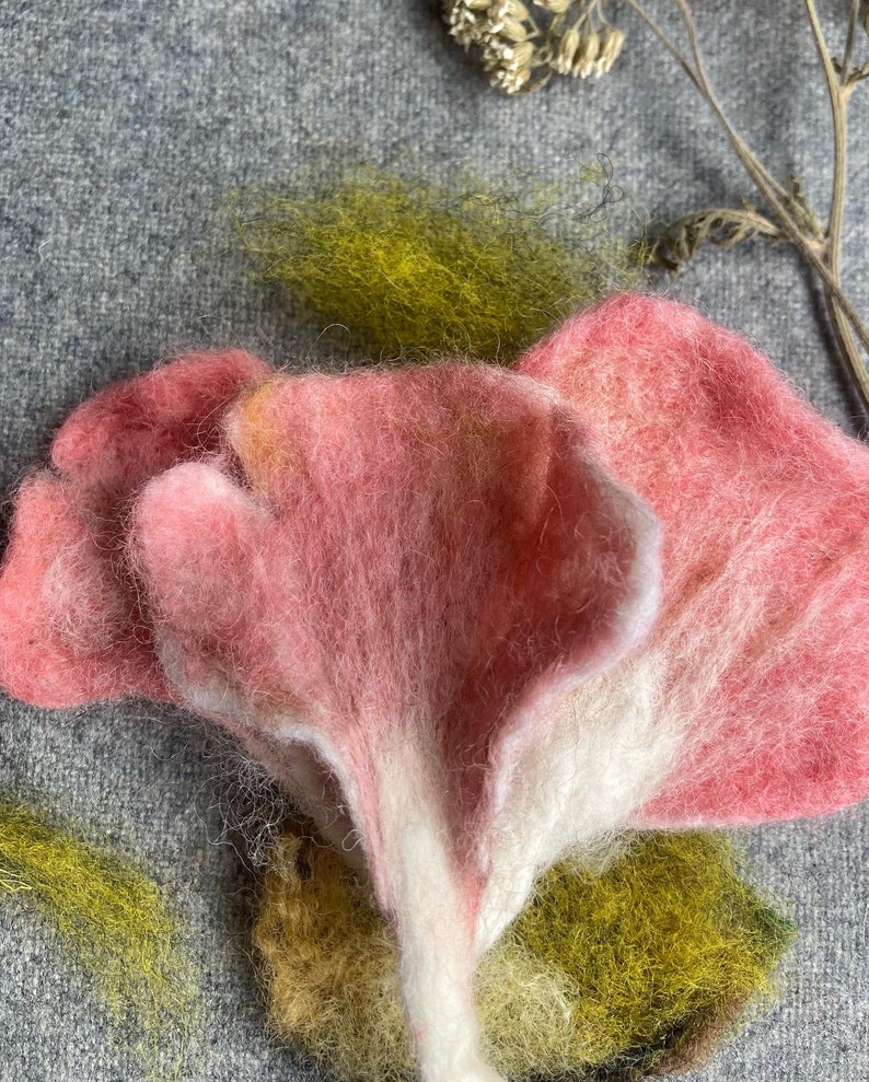 Puede incluir: Un hongo de fieltro rosa y blanco con una superficie texturizada. El hongo est&aacute; hecho de lana y tiene una forma delicada y org&aacute;nica.