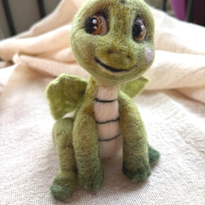 Puede incluir: Una figura de dragón verde de lana fieltrada con grandes ojos marrones, vientre color crema claro con costuras horizontales oscuras, alas pequeñas y un suave rubor rosado en las mejillas. La criatura tiene una superficie texturizada y sonríe.