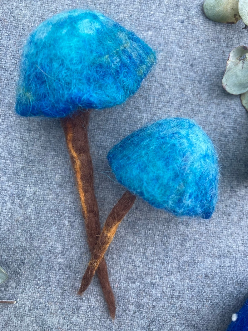 Puede incluir: Dos setas de fieltro azul con tallos marrones. Las setas est&aacute;n hechas de fieltro de lana y tienen una textura peluda.