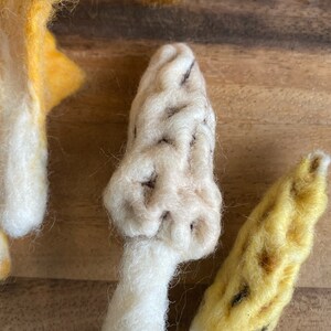 Puede incluir: Dos setas de fieltro, una blanca con detalles marrones y otra amarilla con detalles marrones. Las setas est&aacute;n sobre una superficie de madera.