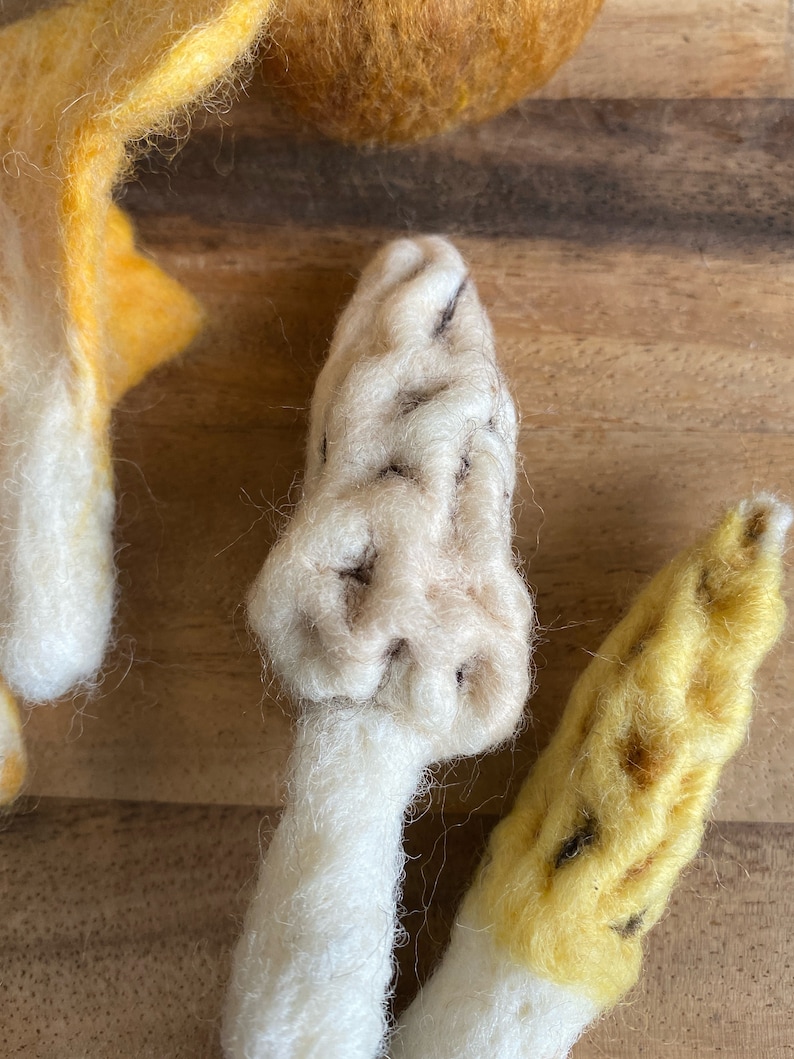 Puede incluir: Dos setas de fieltro, una blanca con detalles marrones y otra amarilla con detalles marrones. Las setas est&aacute;n sobre una superficie de madera.